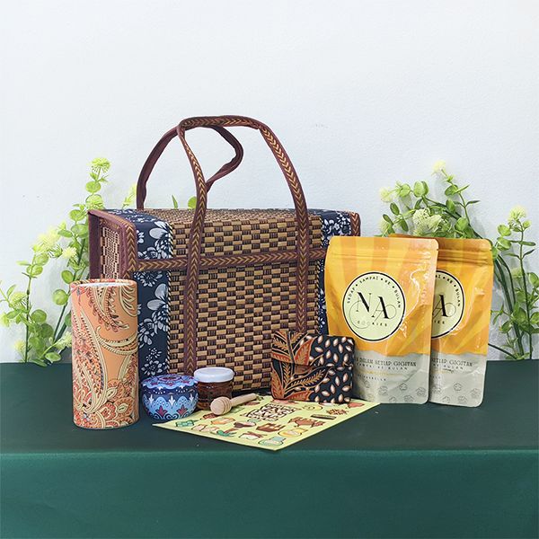 Woven rattan tote basket with leather handles, set out with batik wrapped items, two yellow snack pouches and a printed card.