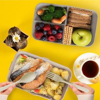 Two-tier bamboo fibre lunch box opened on a yellow table with rice, fish, berries and fruit inside, next to a cup of tea