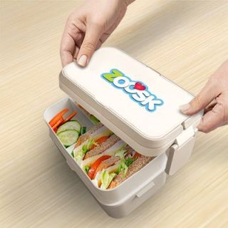 Hands lifting the top tier of the bamboo fibre lunch box to show a salad in the lower tier, with a logo printed on the lid