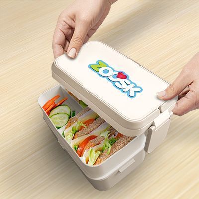 Hands lifting the top tier of the bamboo fibre lunch box to show a salad in the lower tier, with a logo printed on the lid