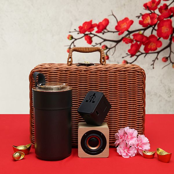 Rattan hamper with a wooden handle, a black thermos and a small wooden speaker, arranged with gold ingots and pink carnations.