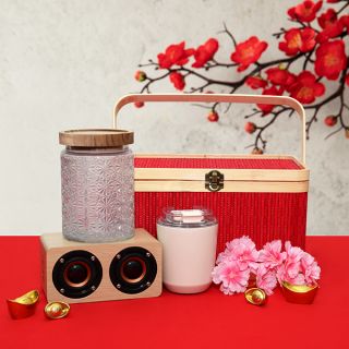 Red tasselled hamper box with a glass canister of pink sweets, a pink tumbler and a wooden twin driver speaker on gold ingots.