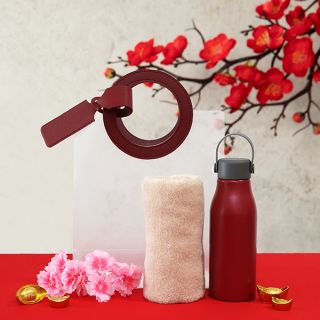 White gift bag with a maroon ring handle and luggage tag, next to a rolled cream towel and a red bottle with a grey carry lid.