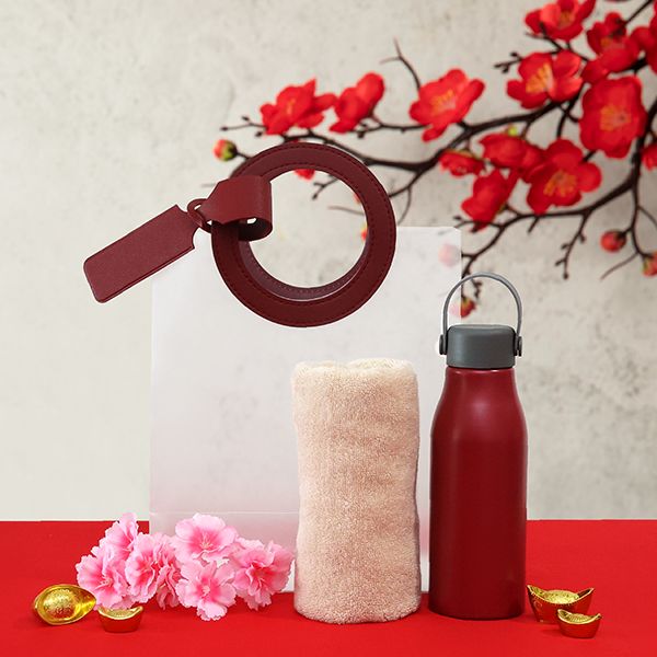 White gift bag with a maroon ring handle and luggage tag, next to a rolled cream towel and a red bottle with a grey carry lid.