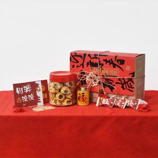 Red patterned gift box with matching cookie canisters and a red packet folder, laid on red cloth for Chinese New Year.