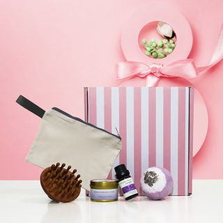 Pink striped box with a cream canvas zip pouch, a round wooden body brush, a small tin, an oil bottle and a lavender bath ball.