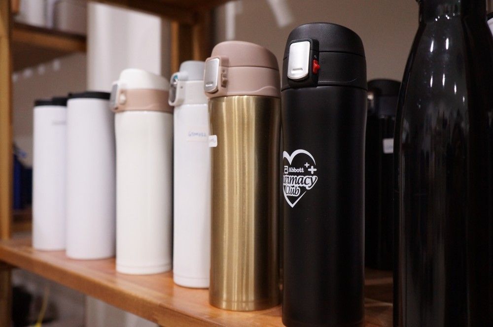 Stainless steel thermos flasks in white, gold and black lined up on a shelf, one engraved with a client's logo